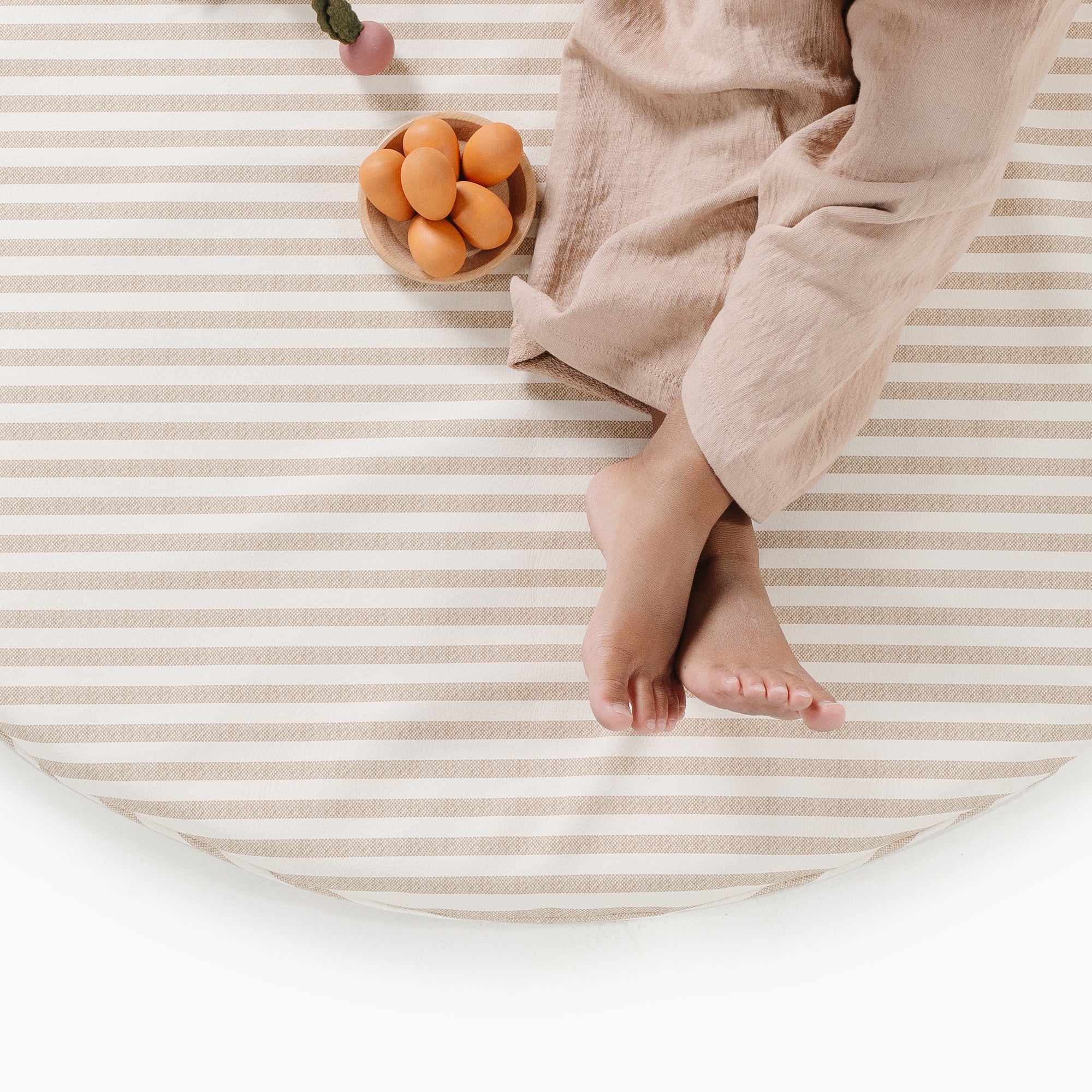 Person wearing beige pants sitting on a striped surface with a small wooden bowl of apricots.
