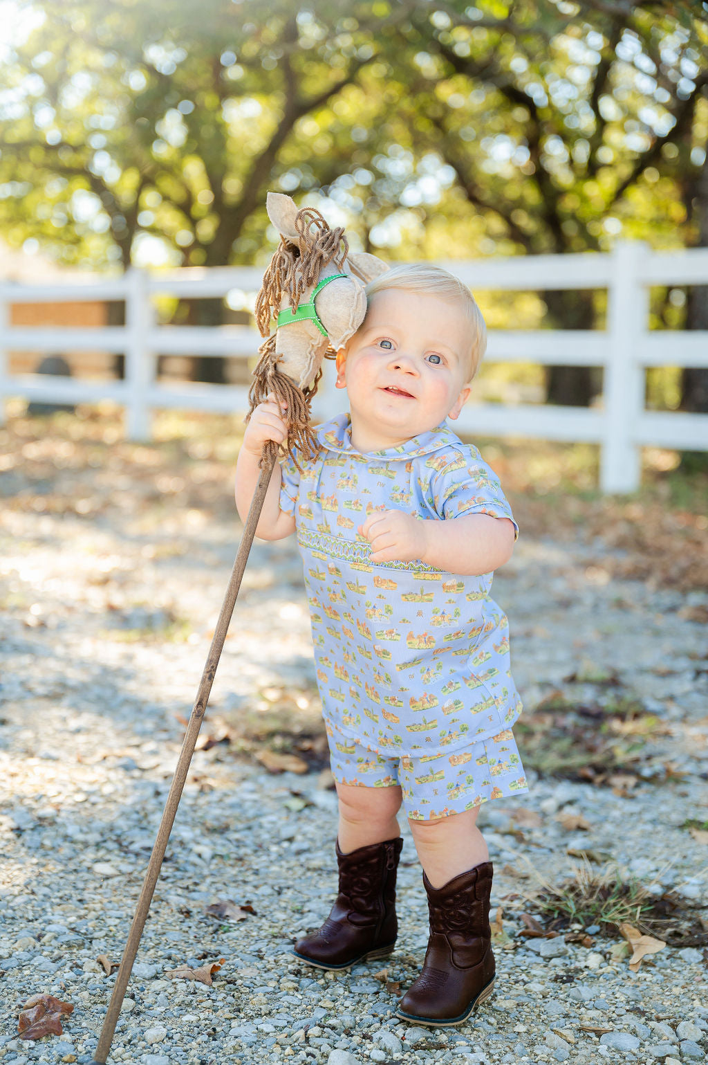 The Smocked Cowboy Toile Samuel Shirt/Shorts Set features a charming cowboy toile print on a blue background, showcasing playful western motifs like horses and boots.