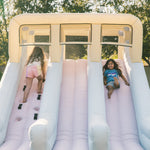 Two children sliding down a pink inflatable slide with a beige top in an outdoor setting.