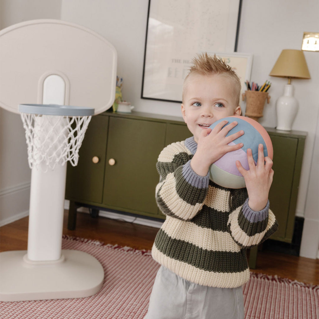 Lifestyle- Child holding a colorful ball in a room with a basketball hoop and furniture.