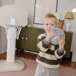 Lifestyle- Child holding a colorful ball in a room with a basketball hoop and furniture.