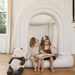 Two young girls sitting on a white inflatable archway, reading a book with a large teddy bear beside them.